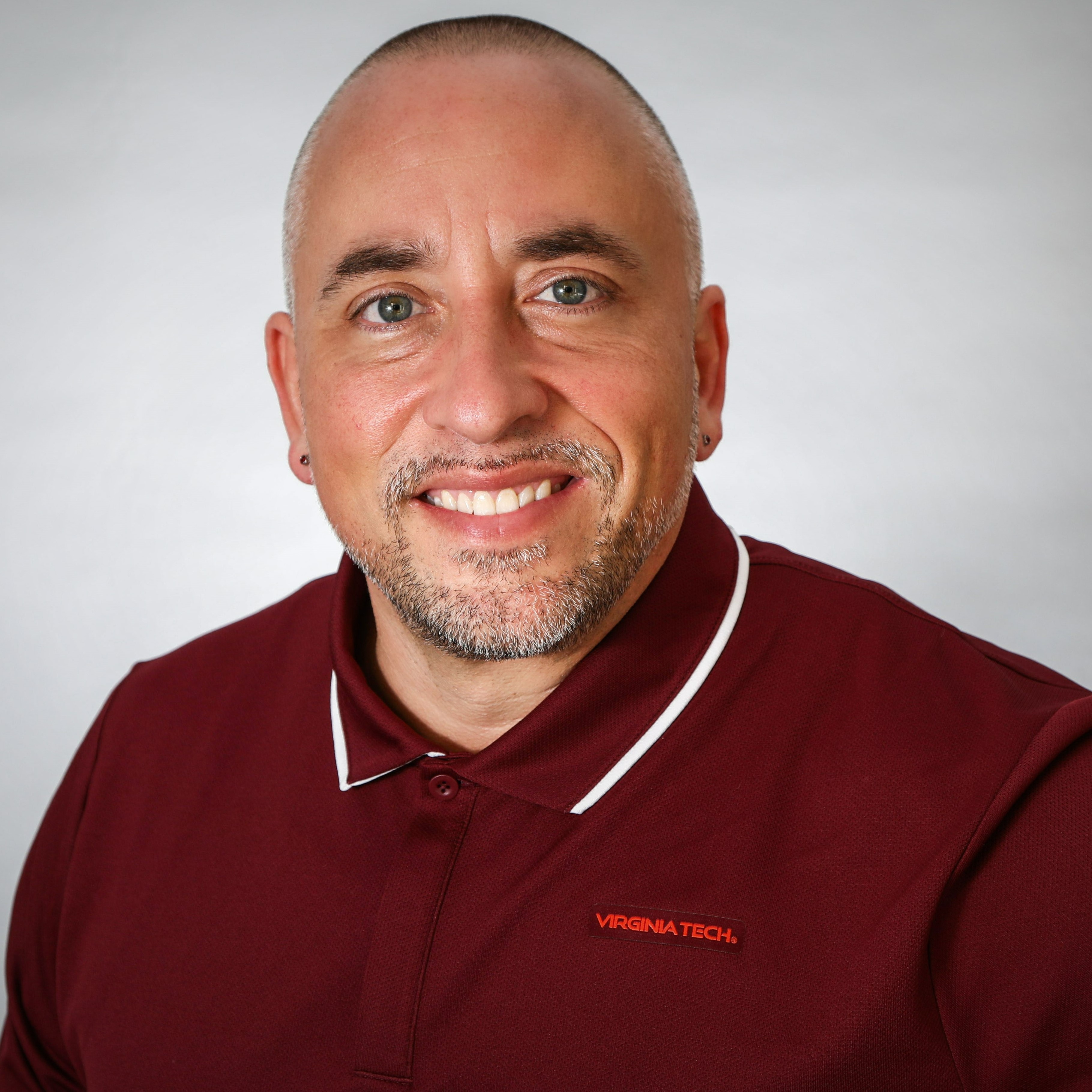 Jon Dance wears a maroon Virginia Tech polo smiling, while against a grey background (chest up)